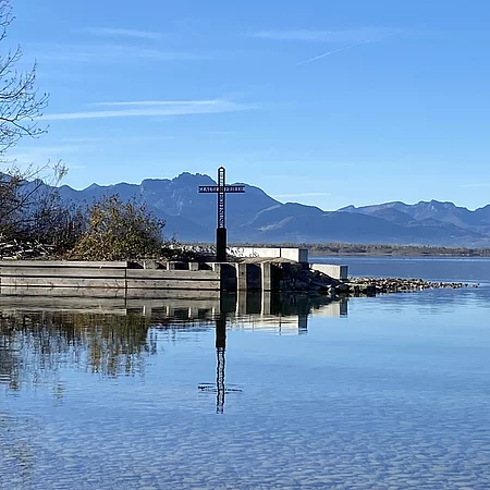 Das Chiemgauer Heimatkreuz auf einer kleinen Landzunge am Thielehafen vor der Kulisse der Chiemgauer Berge